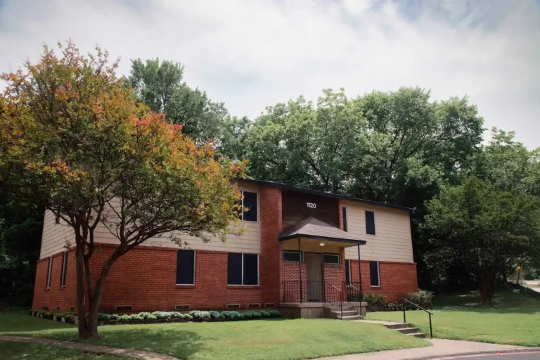 Red brick building with address 1120, surrounded by lush green trees and a manicured lawn under a partly cloudy sky.